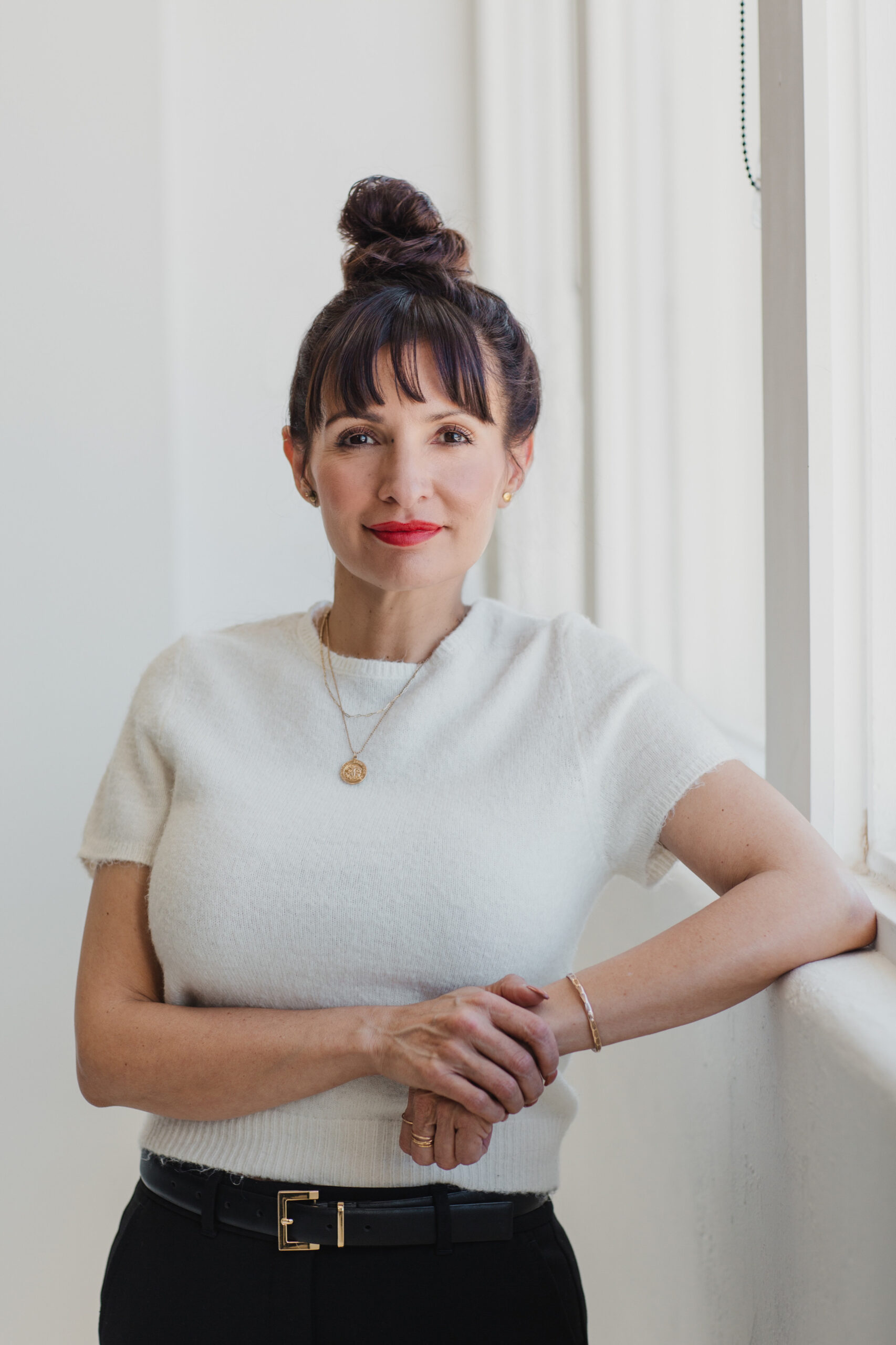 Professional portrait of a woman standing by a sunlit window in a white top and black pants, wearing gold jewelry and red lipstick, photographed by a brand photographer in Albuquerque to visually communicate brand identity and design for a small business.