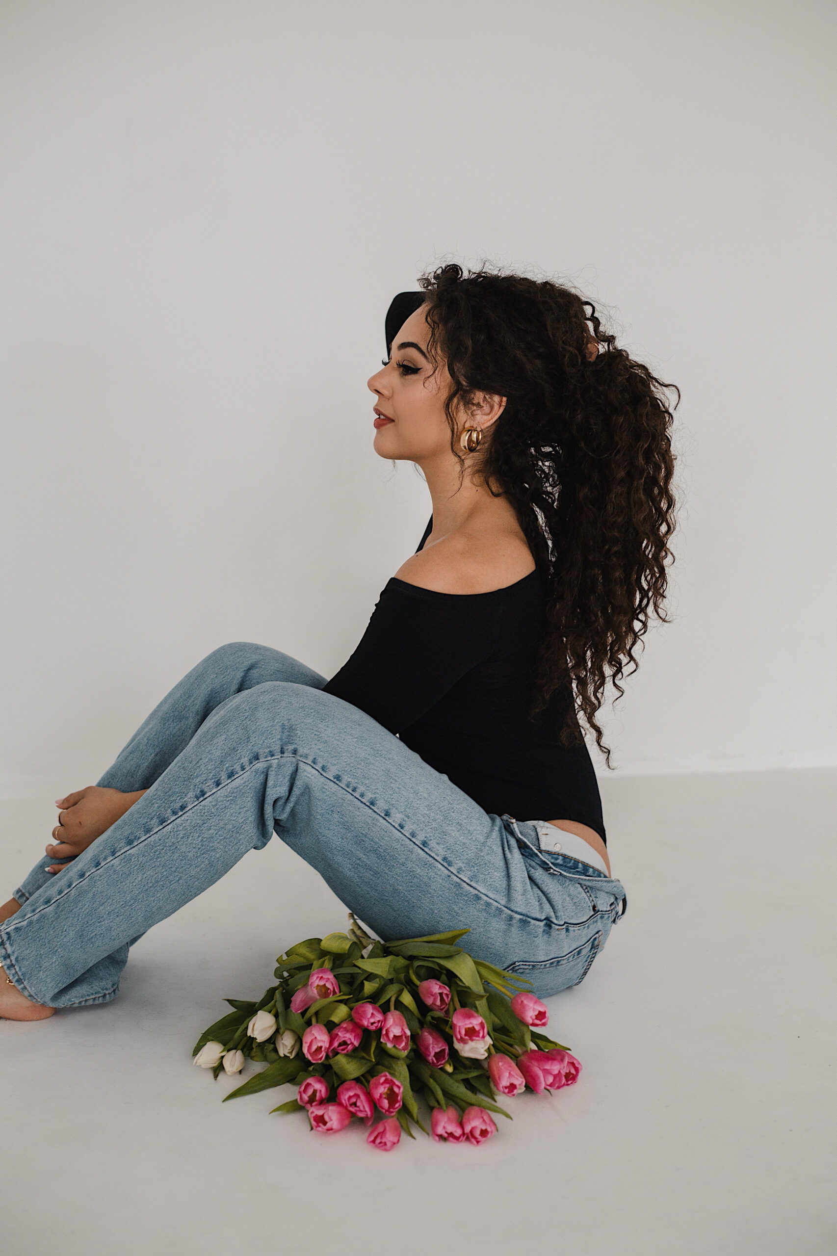 Woman with curly hair in a black off-the-shoulder top and blue jeans sitting on the floor with a bouquet of pink and white tulips, posing in a minimalist studio setting for a branding photoshoot by a brand photographer.