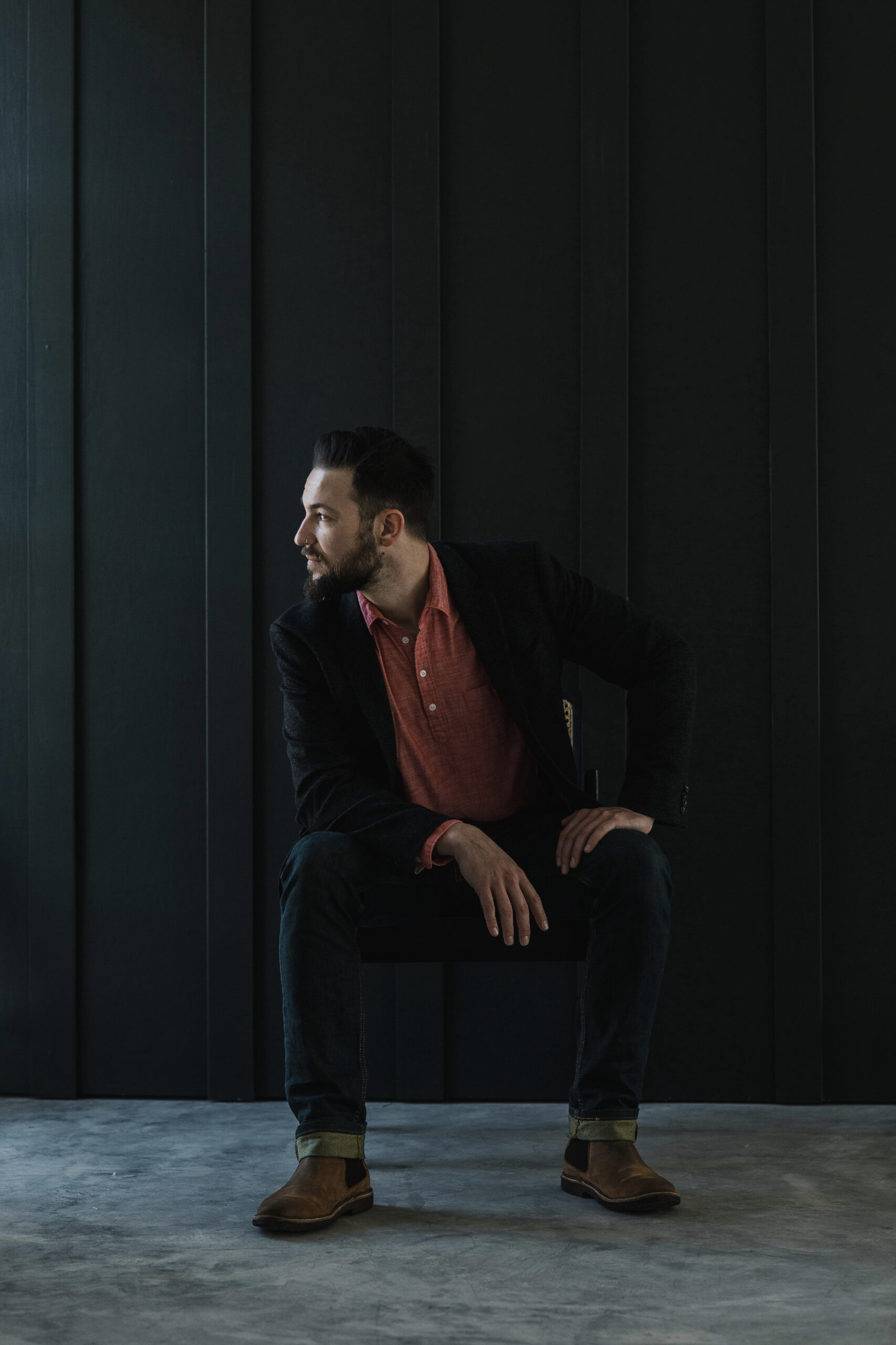Man sitting on a chair in front of a dark paneled wall, wearing a red shirt, dark blazer, jeans, and brown boots, looking to the side, featured in a branding photoshoot by a brand photographer.