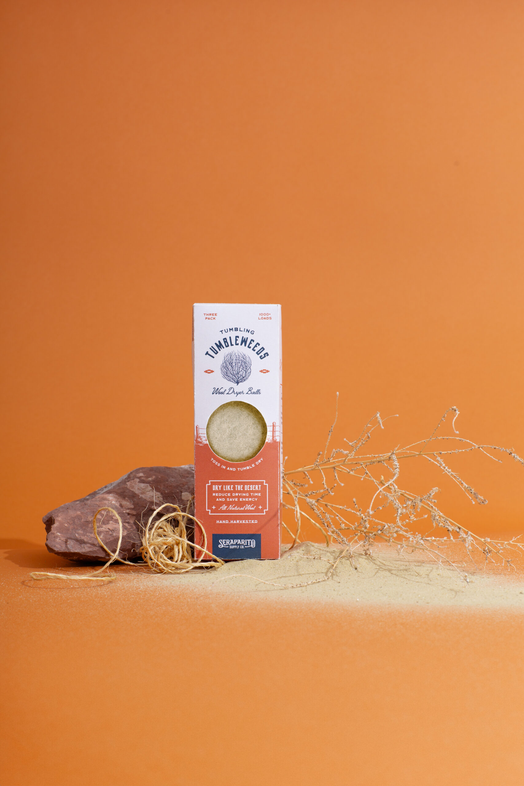 Product packaging for wool dryer balls labeled “Tumbling Tumbleweeds” displayed with desert-themed props on an orange background, creatively styled to emphasize product identity and branding in photography.