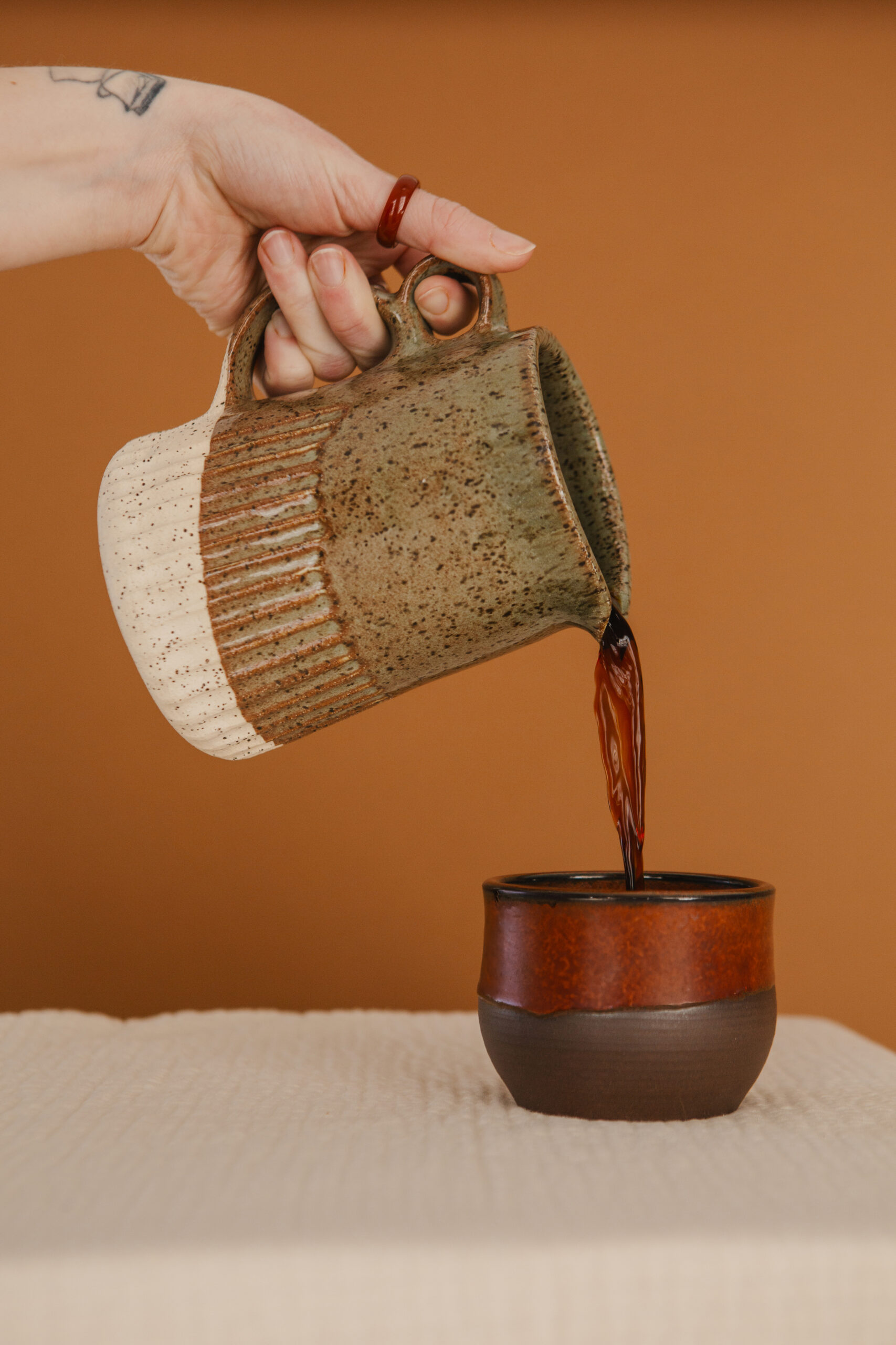 Hand pouring coffee from a speckled ceramic pitcher into a brown handcrafted mug, set against a warm-toned background, styled to evoke a cozy, artisanal atmosphere for custom stock images in small batch product branding.