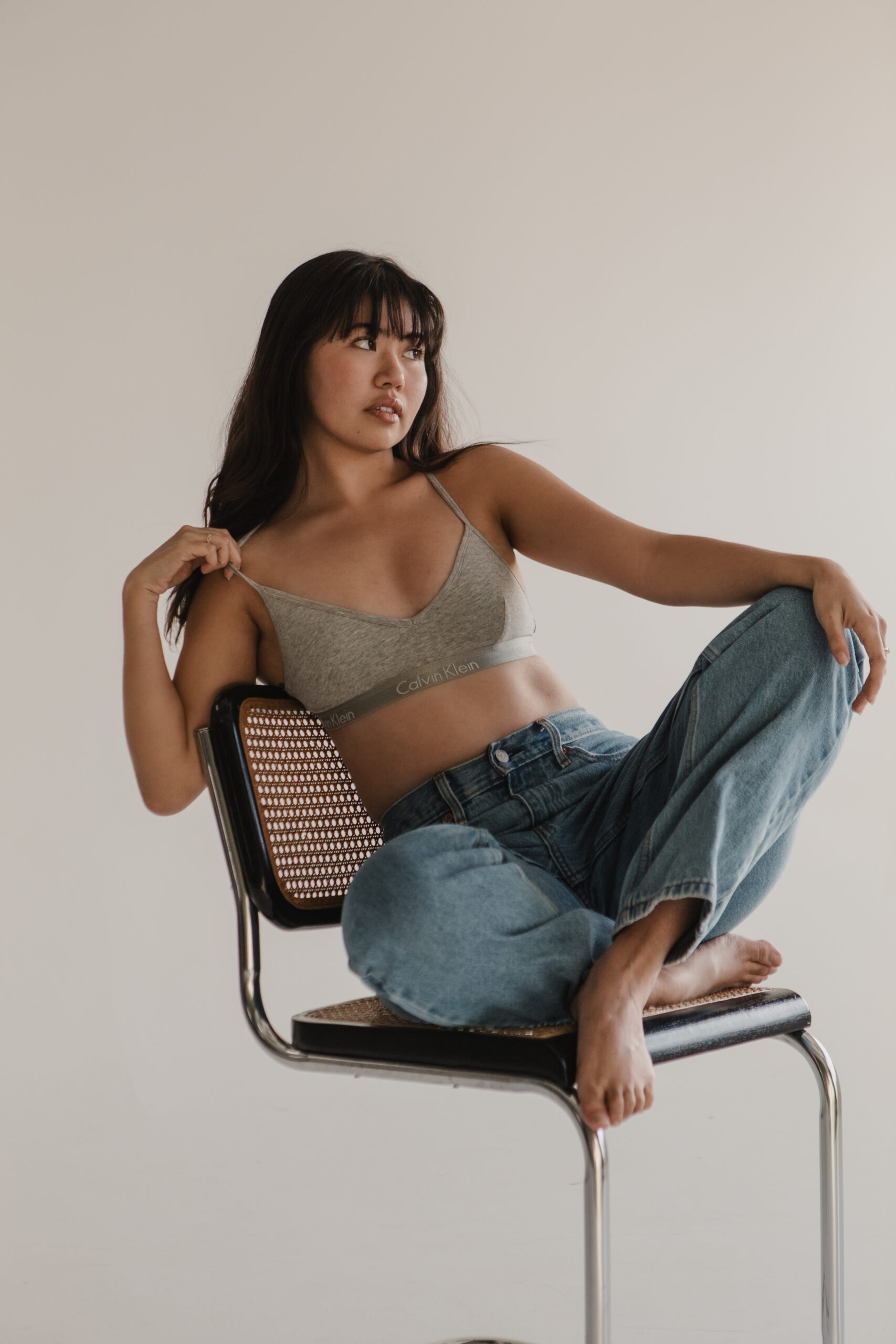 Woman in a gray Calvin Klein bralette and relaxed-fit jeans sitting barefoot on a woven-back chair, looking off to the side, photographed by a branding photographer for modern casual headshots.