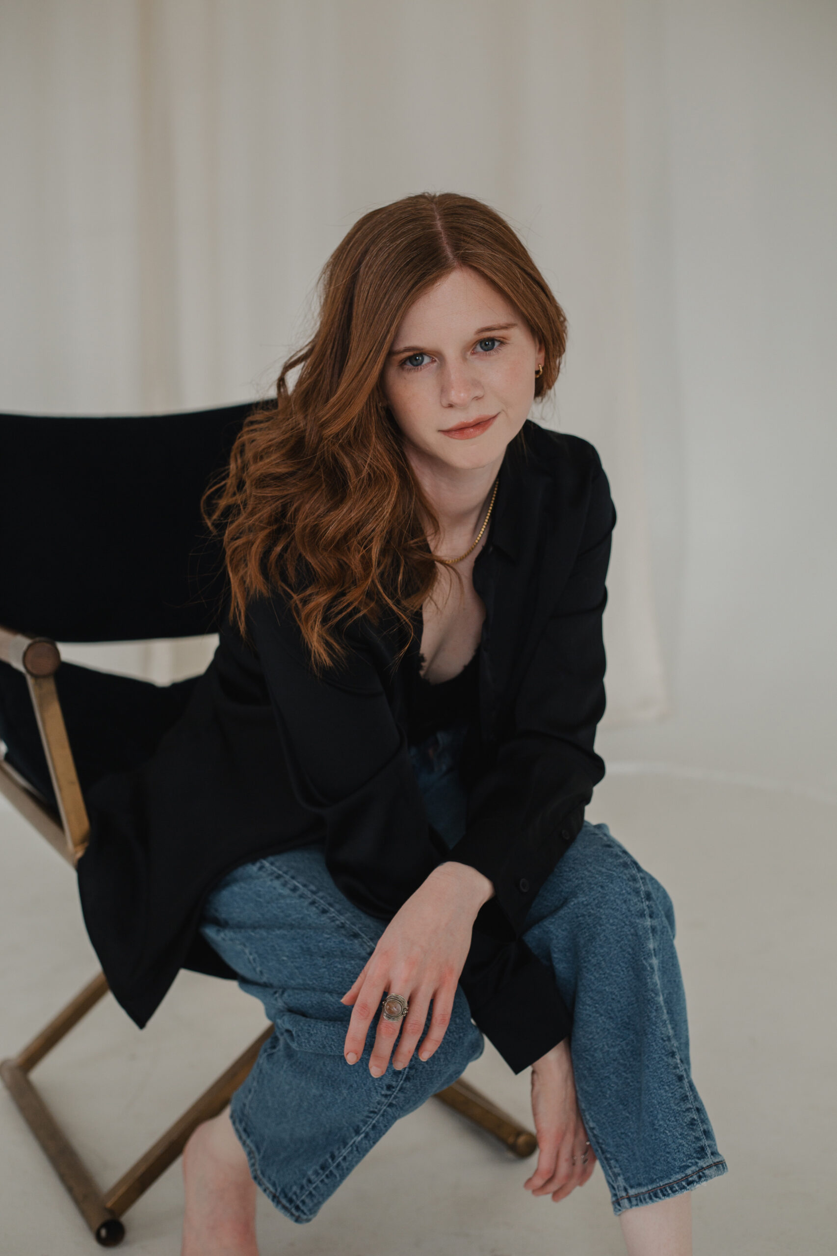 Woman with long red hair seated on a director-style chair, wearing a loose black button-up shirt and relaxed blue jeans, leaning forward with a confident gaze—photographed by a branding photographer to convey professionalism with a modern, creative edge.