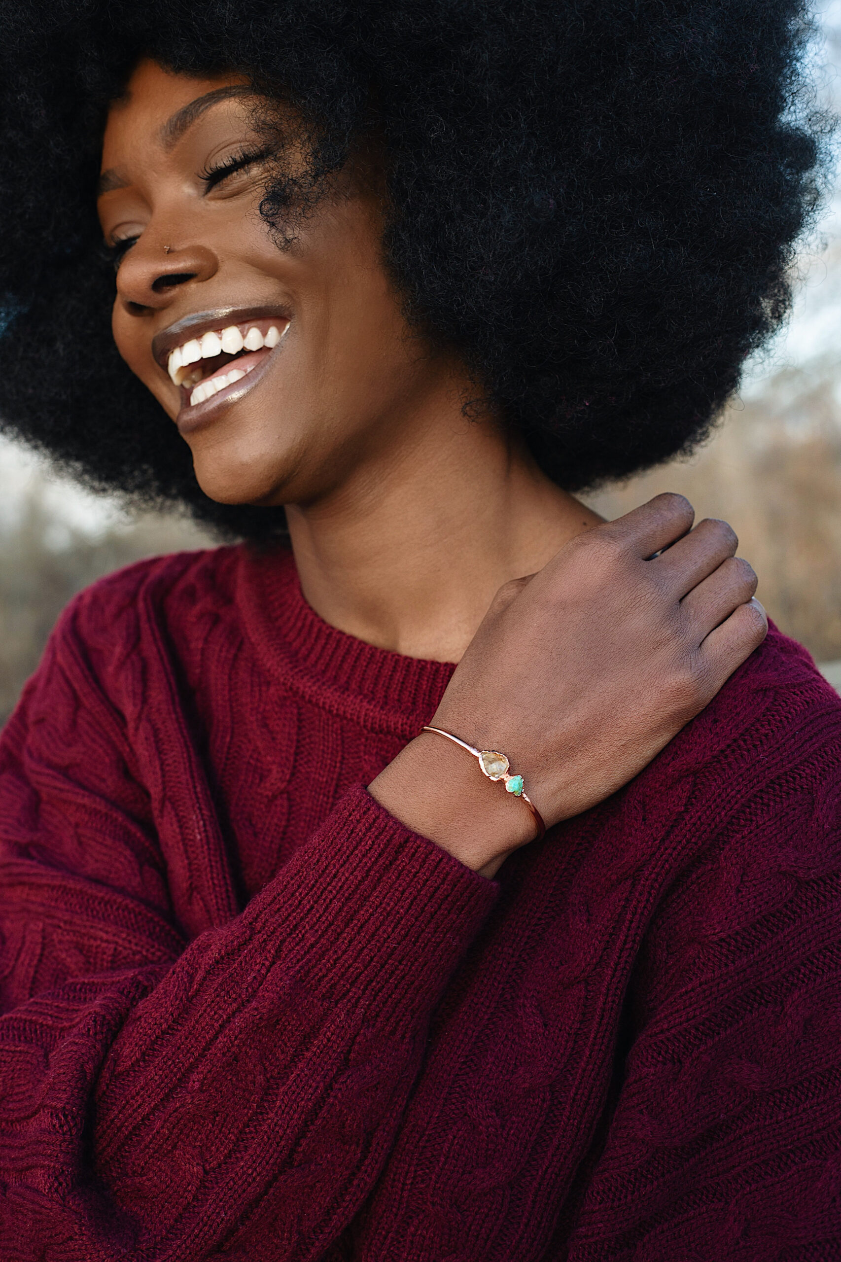 Close-up of a woman smiling and wearing a gemstone bracelet while dressed in a burgundy cable-knit sweater, captured outdoors during a headshots studio session by a branding photographer.