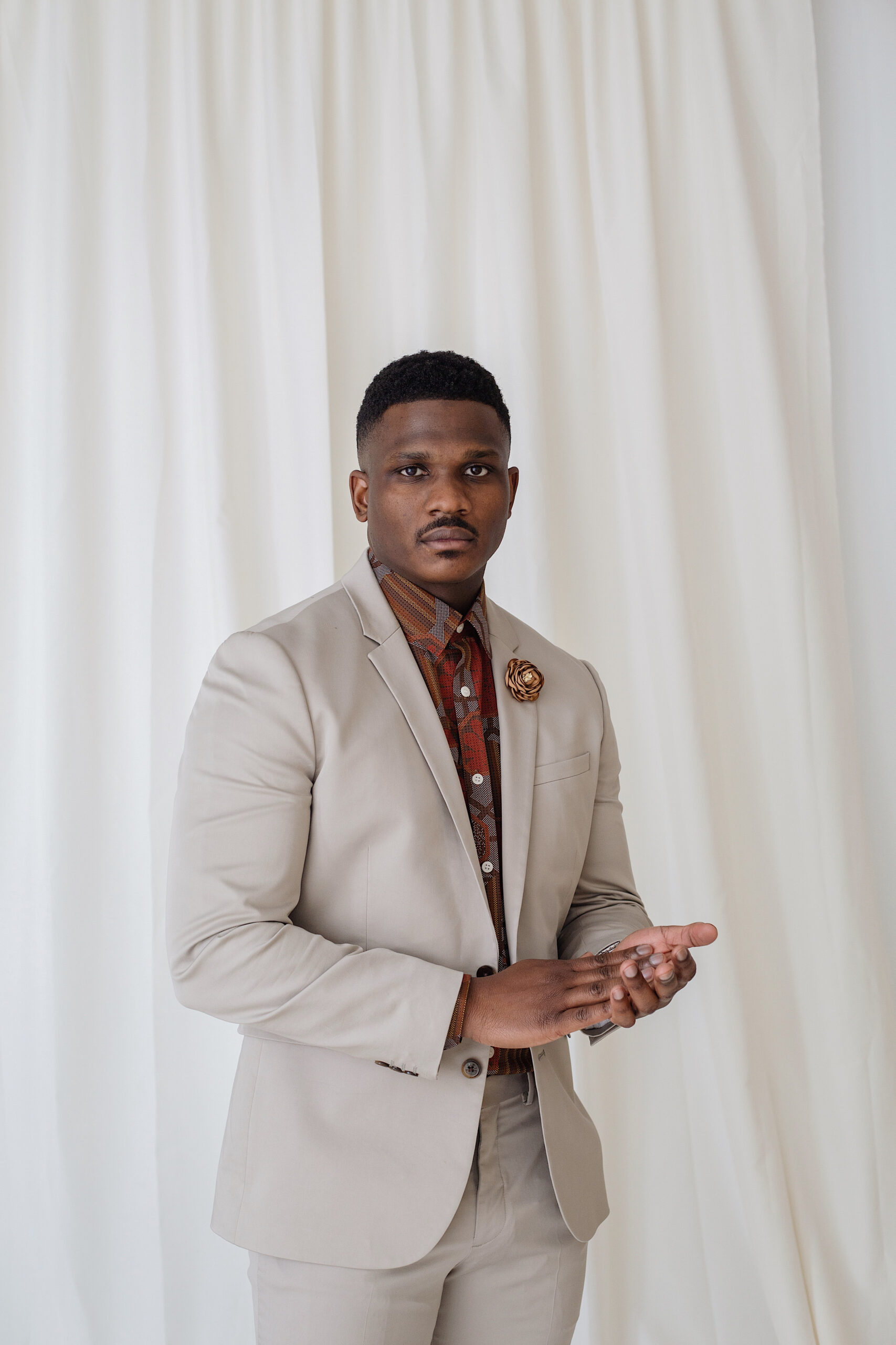 Man in a light beige suit with a patterned brown shirt and floral lapel pin posing in front of white curtains, captured during a headshots studio session by a branding photographer.